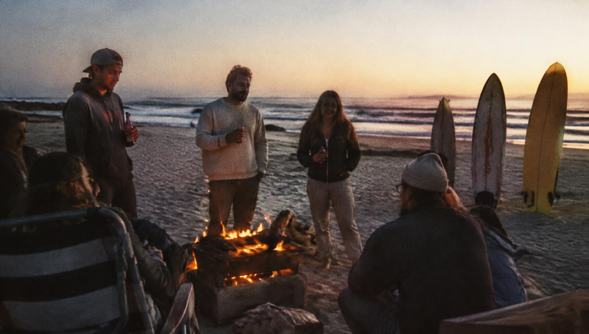 Friends around a beach braai at dusk with surfboards in the sand